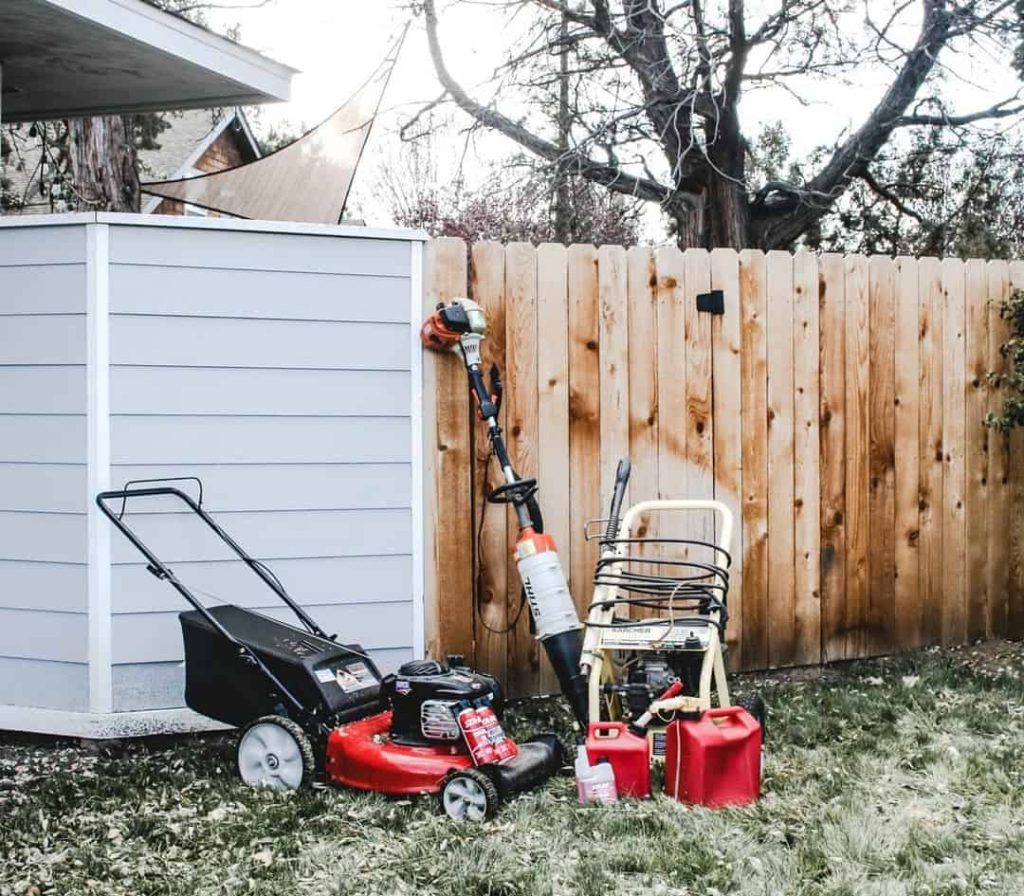 Push mower, power washer, gas cans and leaf blower sitting next a privacy fence.