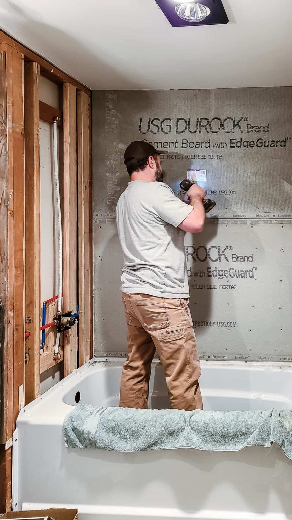 A man is securing Durock cement board an unfinished bathroom with a bathtub with a drill
