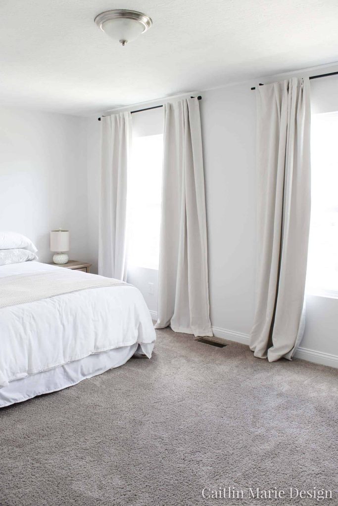 Cameo White from Behr Paint is a great pearly white paint color that looks great painted on the walls in this bedroom from Caitlin Mair Deisgn with gray carpeting, cream linen curtains and a bed with white bedding 