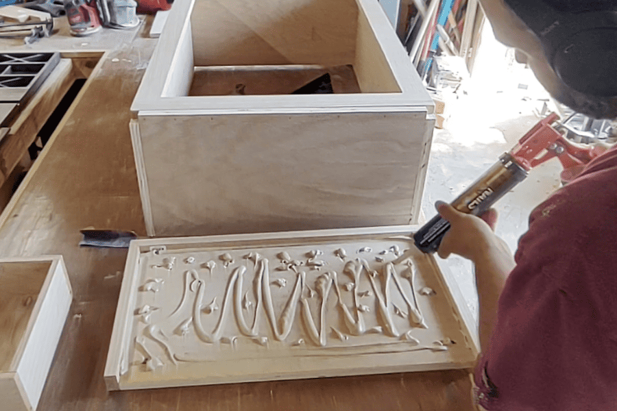 A man applies adhesive to plywood top before attaching to the top of the DIY trash can cabinet
