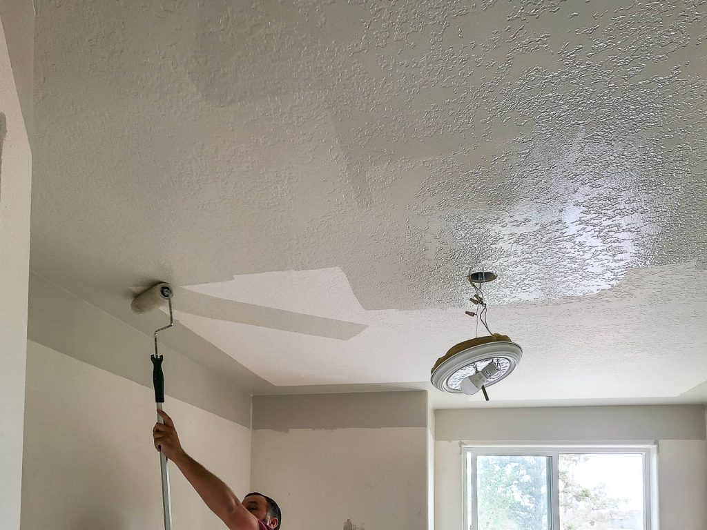 Man painting the ceiling with overhead light fixture hanging down away from the ceiling.