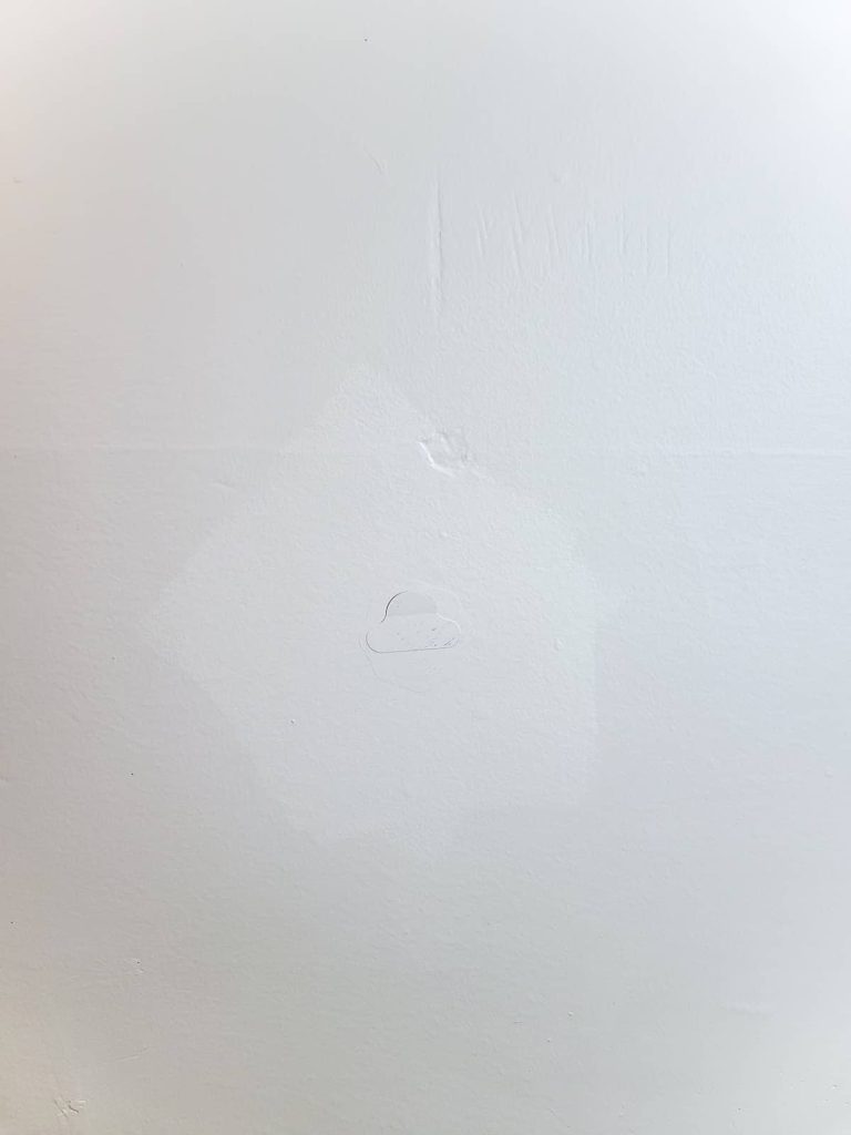 A close up image of a patch of wet paint on a wall, with a small sticker in the center, covering a hole in the drywall.