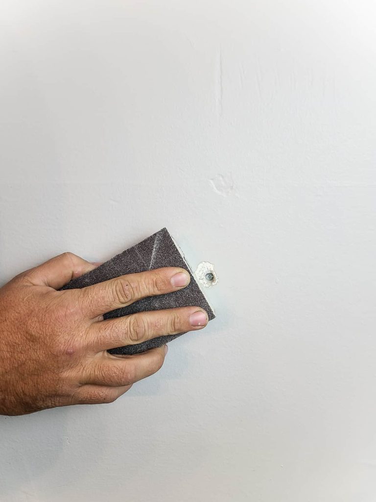 An image shows a person using a sanding block to sand the wall around a small hole.