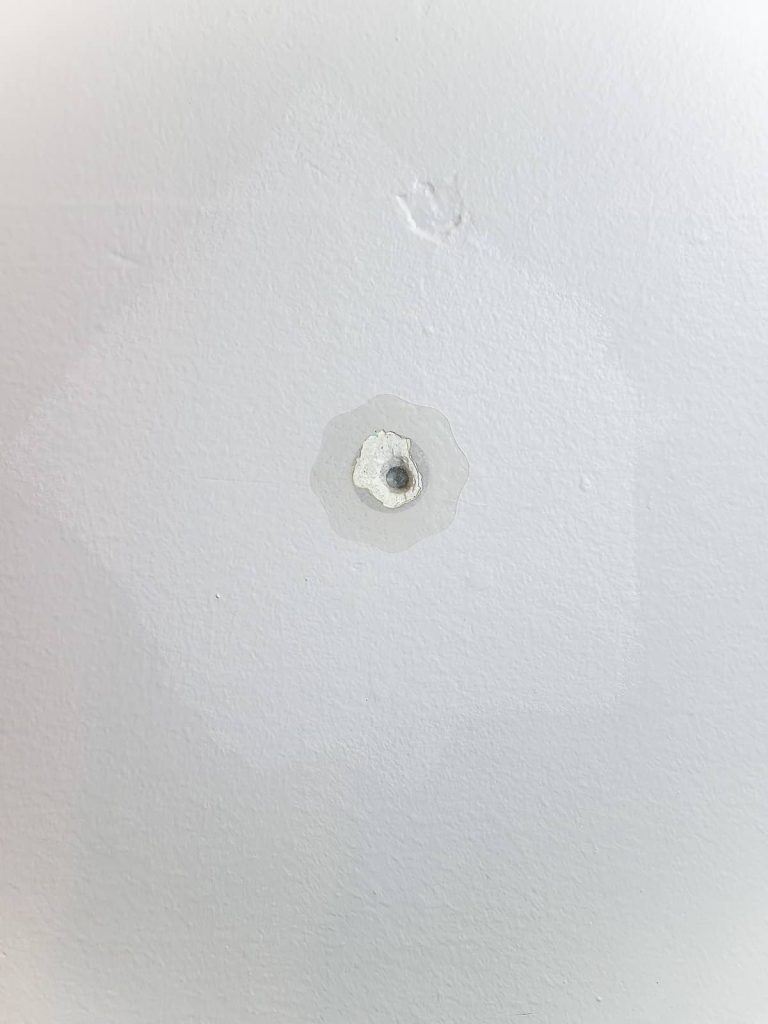 A closeup image of a swatch of fresh paint on the wall, which is lighter than the surrounding paint. In the center is a small hole, covered by a transparent DAP repair patch.