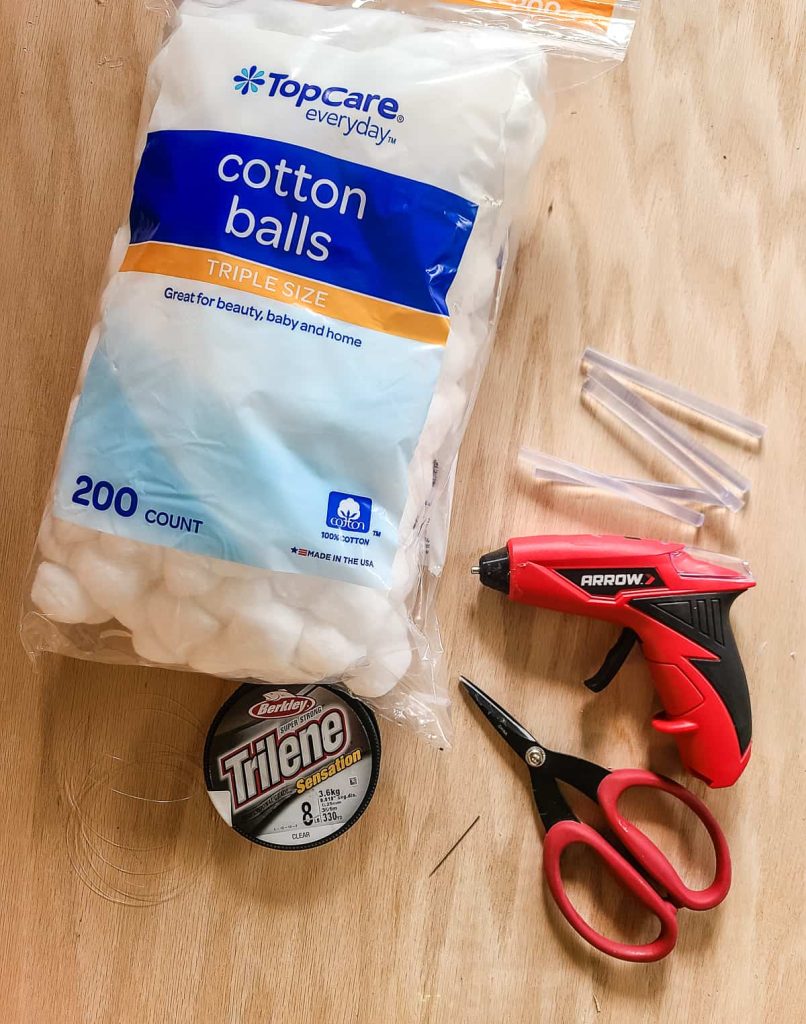 Supplies needed to make cotton ball snowball wreath laying on flat surface with bag of white cotton balls, fishing line, needle, scissors, hot glue gun and hot glue sticksw