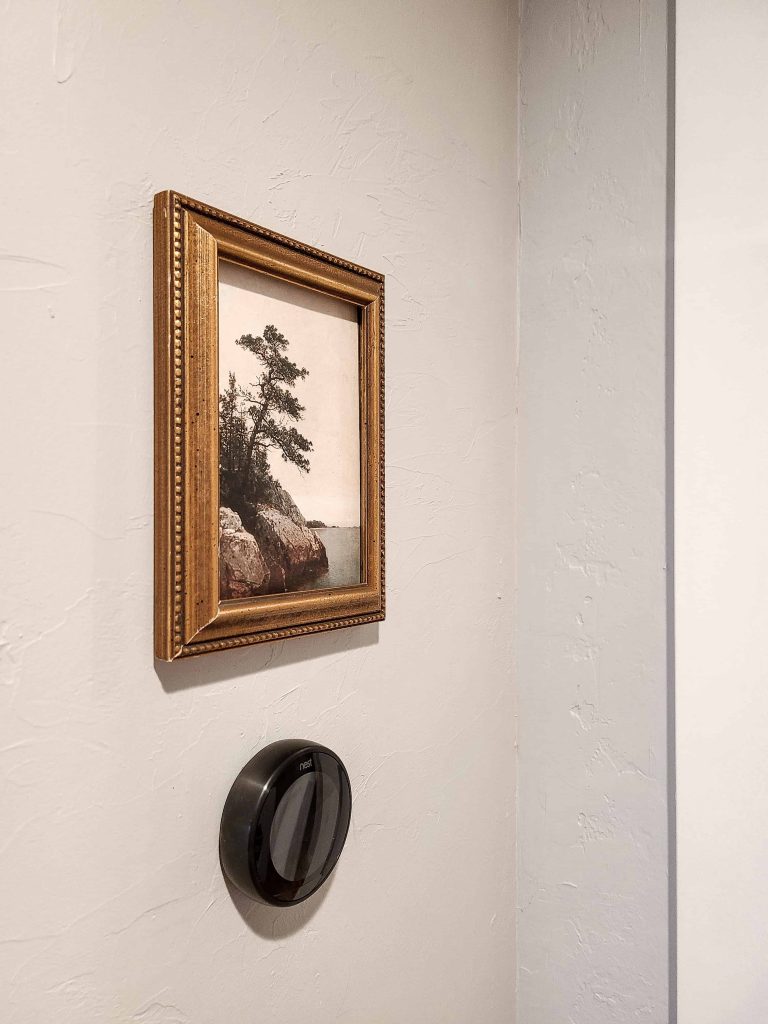 The print made to look like a painting hung up on the wall above a thermostat in a hallway 