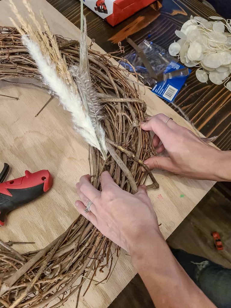 A photo of a person's hand attaching a pampas grass into a grapevine wreath.