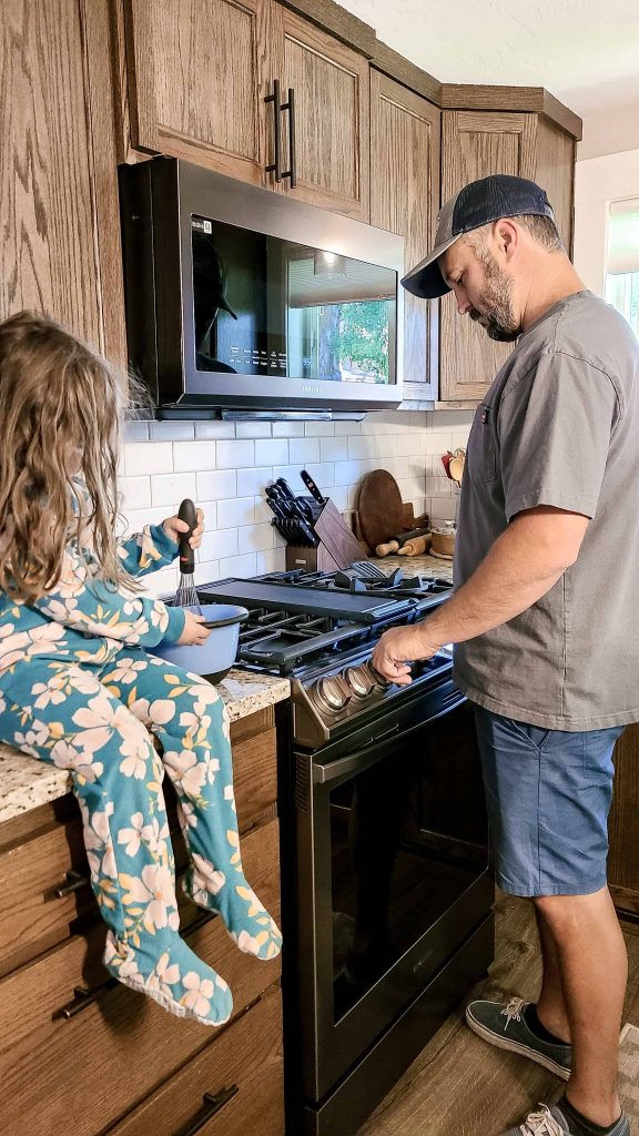 A girl sits at a table beside the Samsung Bespoke Gas Range as the man stands and adjusts one of the knobs.