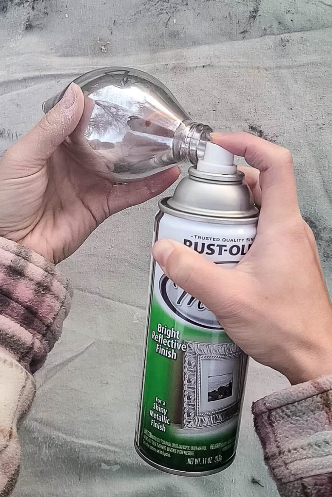 The person sprays a coat of metallic spray paint onto the surface of a mercury glass ornament.
