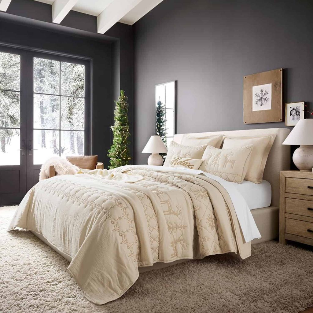 beige colored Christmas themed quick on bed with embroidered designs for reindeer, trees and snowflakes in a room painted with dark gray walls with a small christmas tree in the corner