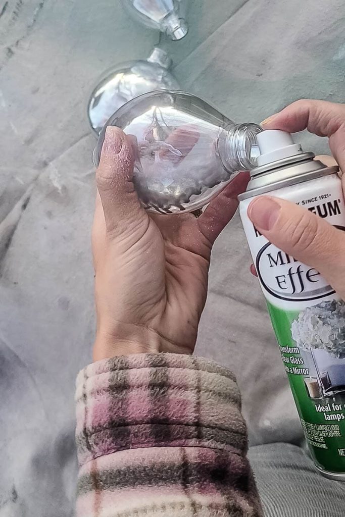 A person sprays the inside of an ornament with one or two small spritzes of mirror spray paint while holding it outside.