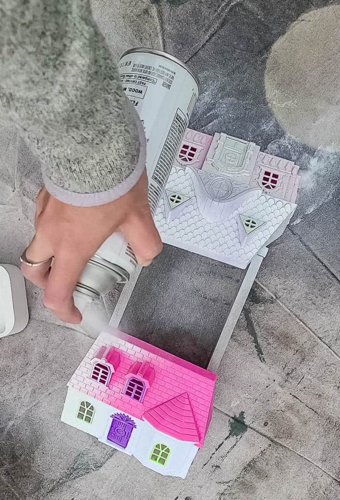 A person is spraying a spray paint onto a cute doll house.