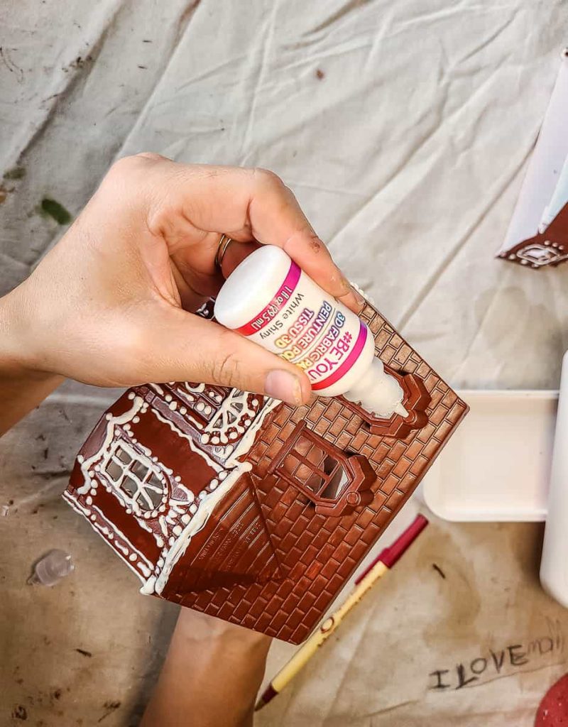 Decorating a gingerbread house with white puffy paint, adding a delightful snowy texture for a festive touch.
