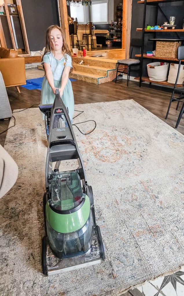 In the photo, a girl is seen using a carpet vacuum cleaner to clean the carpet.