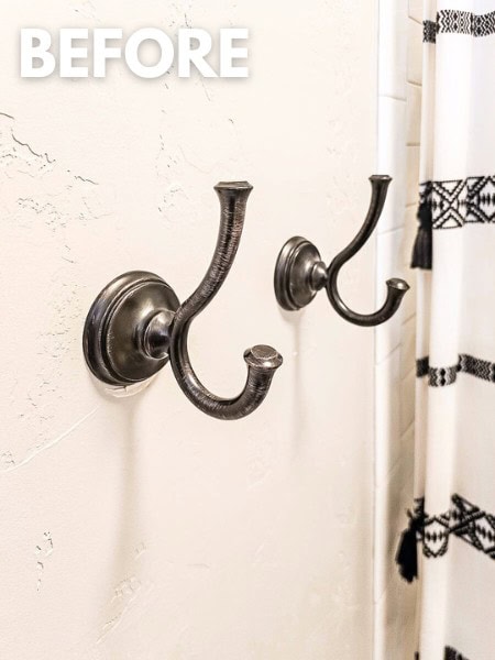 Close up of oil rubbed bronze towel hooks in bathroom in a gray painted wall. Towel hooks are from Delta with the Cassidy line. Image has text overlay that says "before"