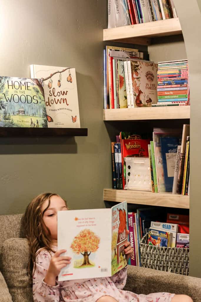 Organized children's books on bookshelves with little girl sitting in nook and reading a picture book
