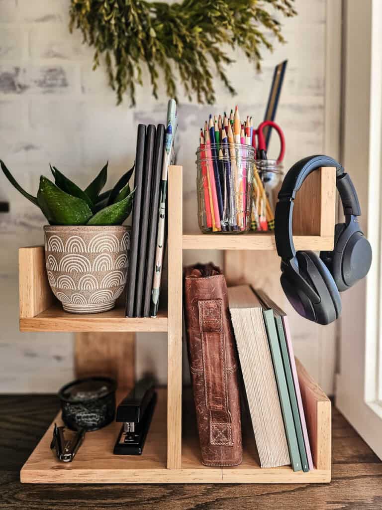 A photo of the finished small bookshelf on a desk, holding books, paper files, journals, pens, pencils, plants, and other office supplies.