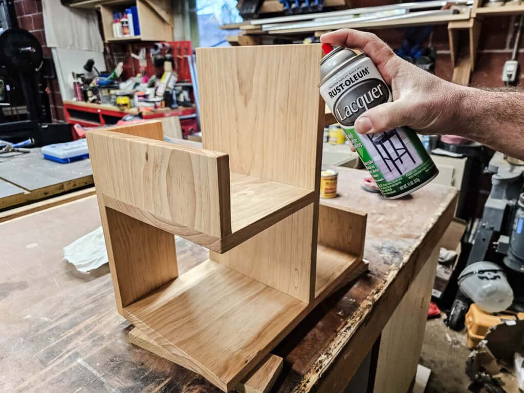 A photo of a man applying Rust-Oleum lacquer to the small bookshelf, using a spray can or brush to coat the wood for a durable, glossy finish, enhancing its appearance and protection.