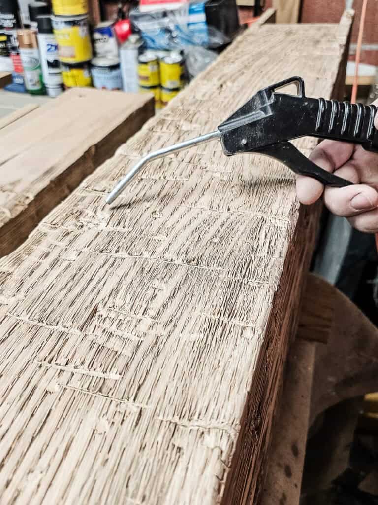 A photo of a man using compressed air to blow sanding dust off the wood, preparing it for staining.