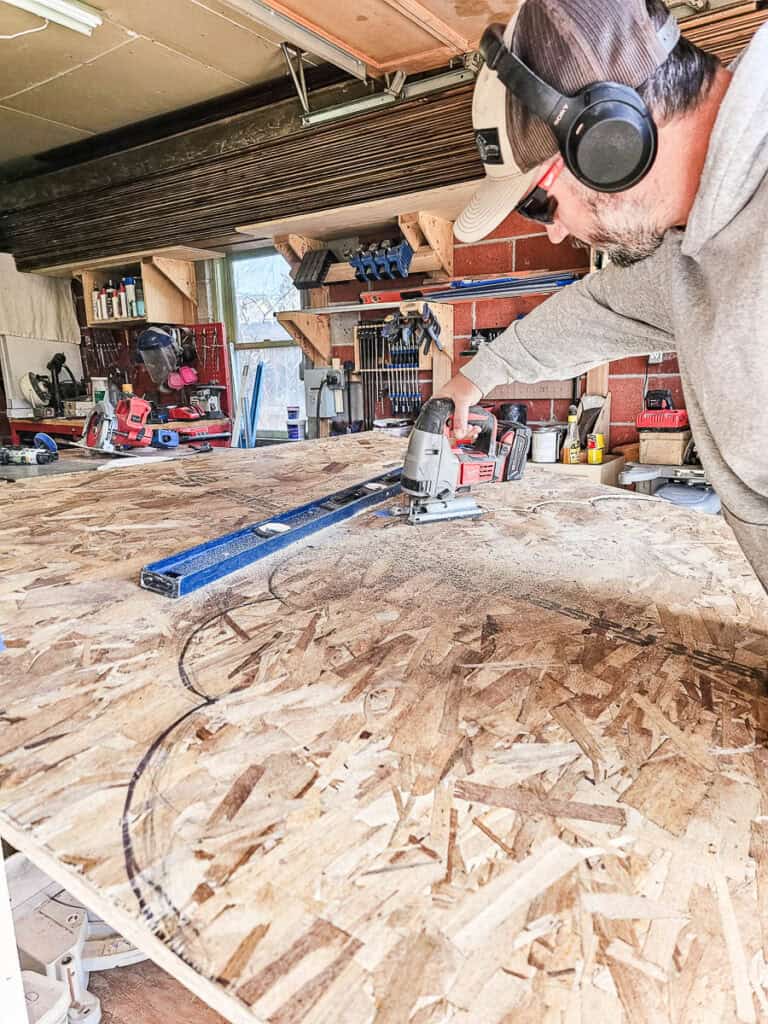 A man is cutting a piece of wood along a drawn cloud-shaped outline  with a jigsaw
