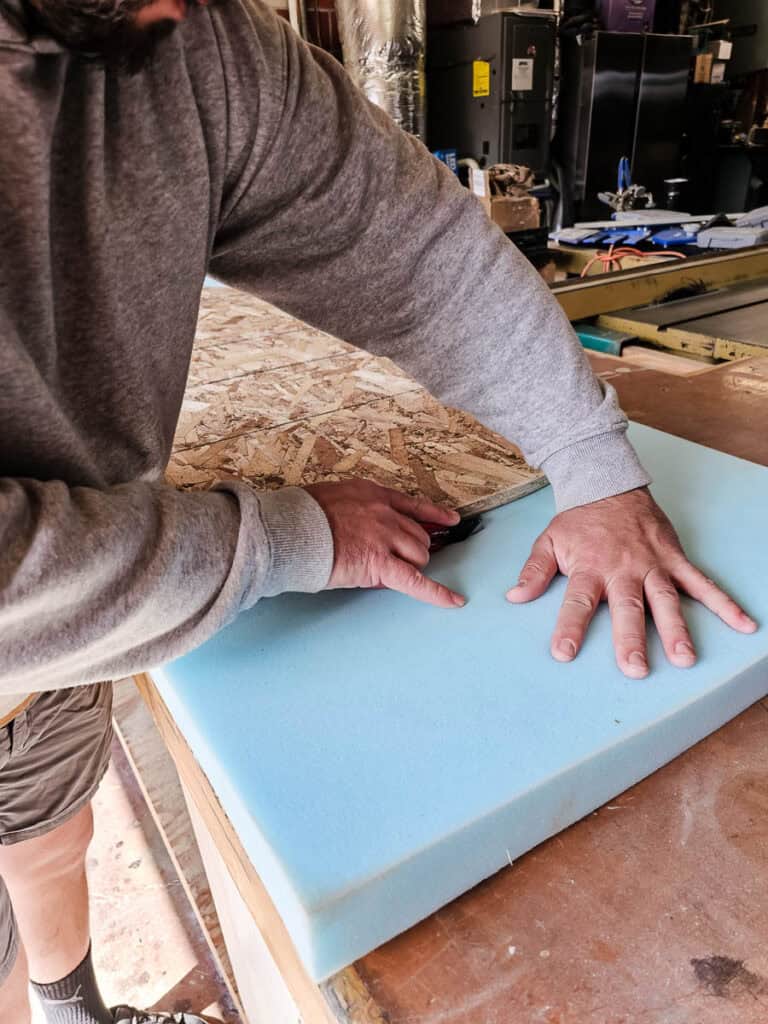 The man is cutting the foam into a with a utility knife for an upholstered headboard 