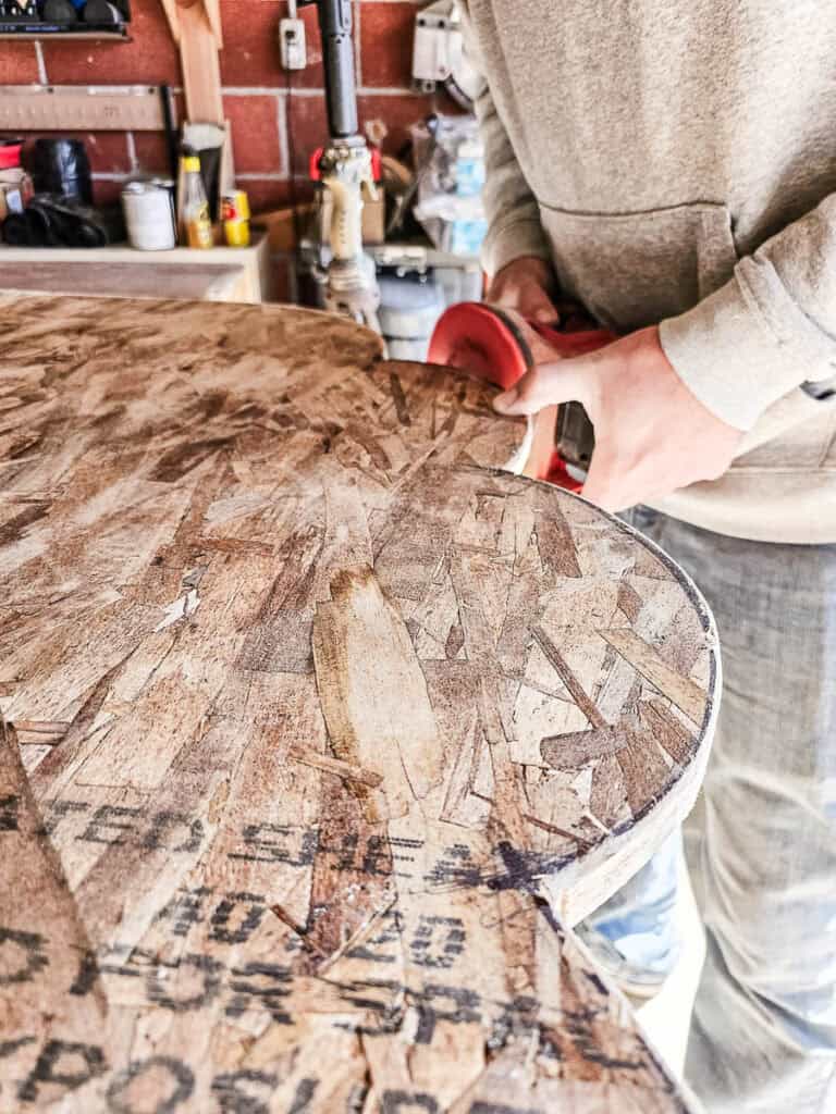 A man continues sanding, using an orbital sander on the edges of the cloud cut from OSB
