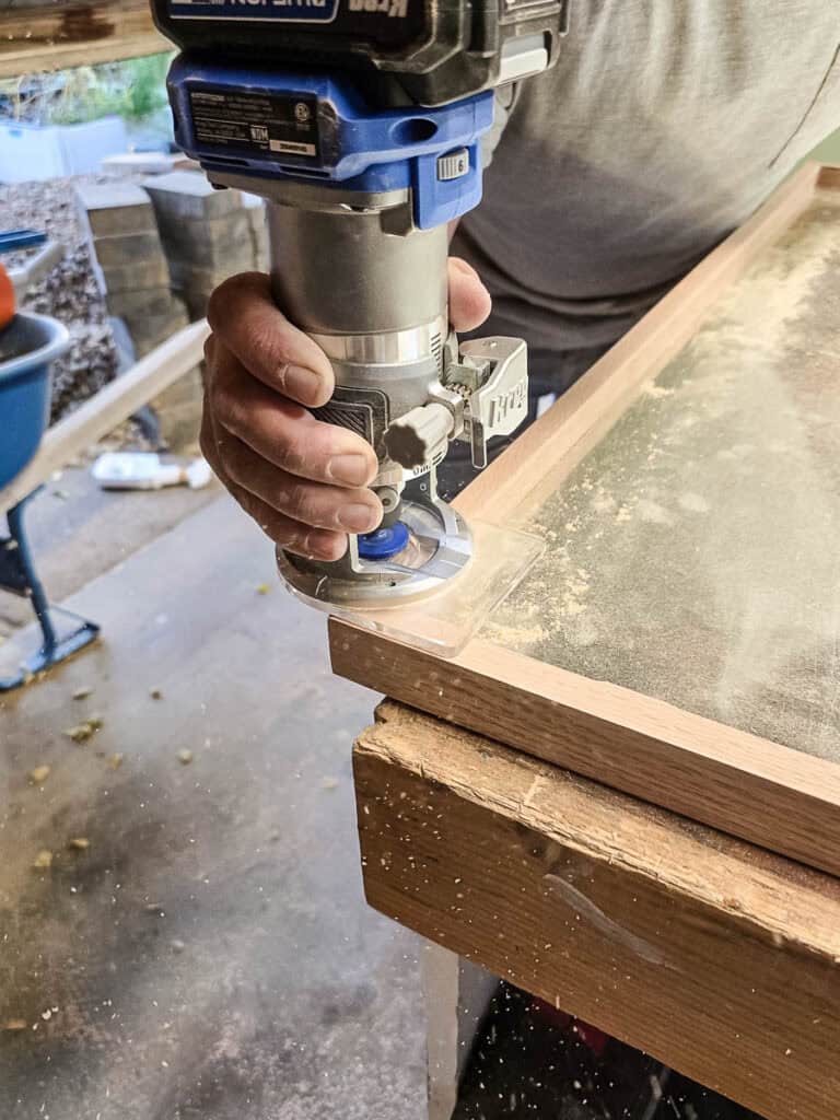 A man using a trim router with a chamfer bit to smooth and angle the frame edges.