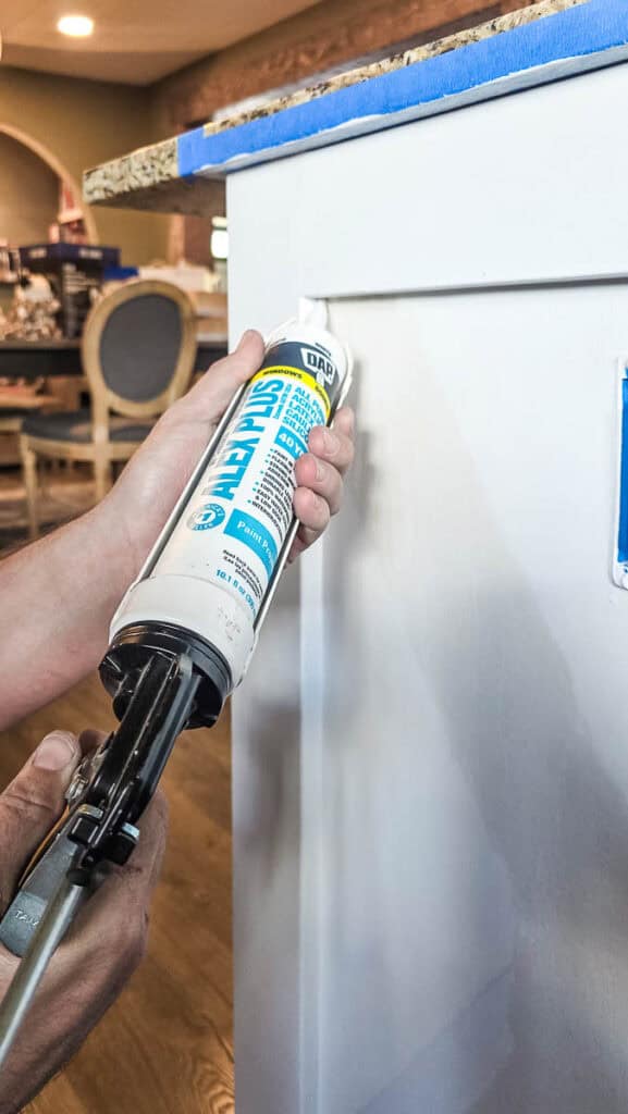 Applying paintable acrylic latex caulk to the seams of a primed kitchen island to fill gaps before painting for a smooth, seamless finish.