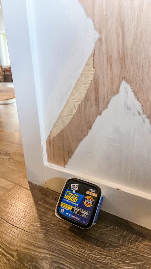 Close-up of a kitchen island with paint removed to expose bare wood and wood filler placed nearby, showing surface repair before priming and painting.