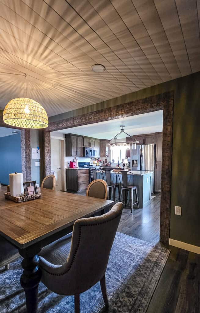 Wide view of a completed kitchen renovation featuring a freshly painted gray-green kitchen island, granite countertops, wood cabinets, and an open dining area with farmhouse-style décor.