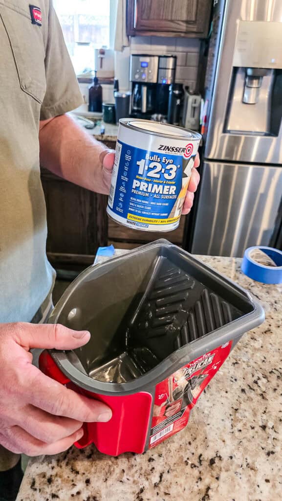 Person holding a can of bonding primer and a paint tray in a kitchen, preparing to prime a kitchen island before painting for better paint adhesion and durability.