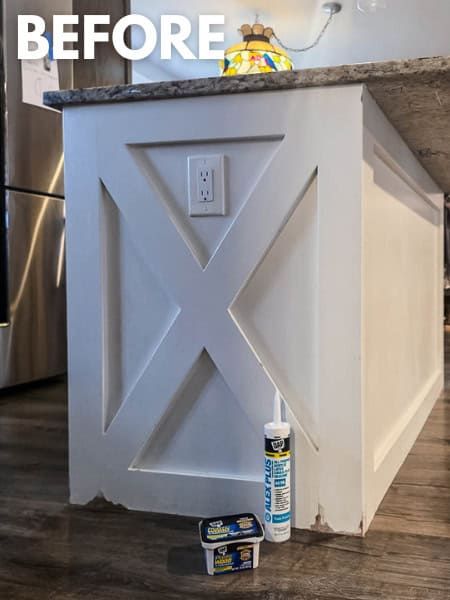 Close-up of a kitchen island before painting, featuring white decorative trim with an X-pattern, granite countertop, and visible chipped paint and caulk repairs before refinishing.