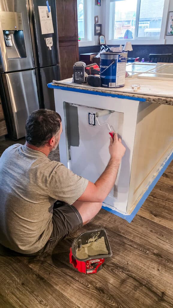 Person using a foam roller to apply the first coat of cabinet paint to a primed kitchen island, creating a smooth, even finish during a DIY kitchen makeover.