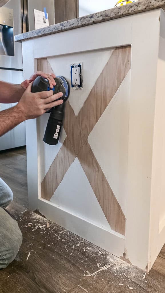 Person using an orbital sander on a kitchen island with decorative X-pattern trim, smoothing the surface before priming and painting for a durable finish.