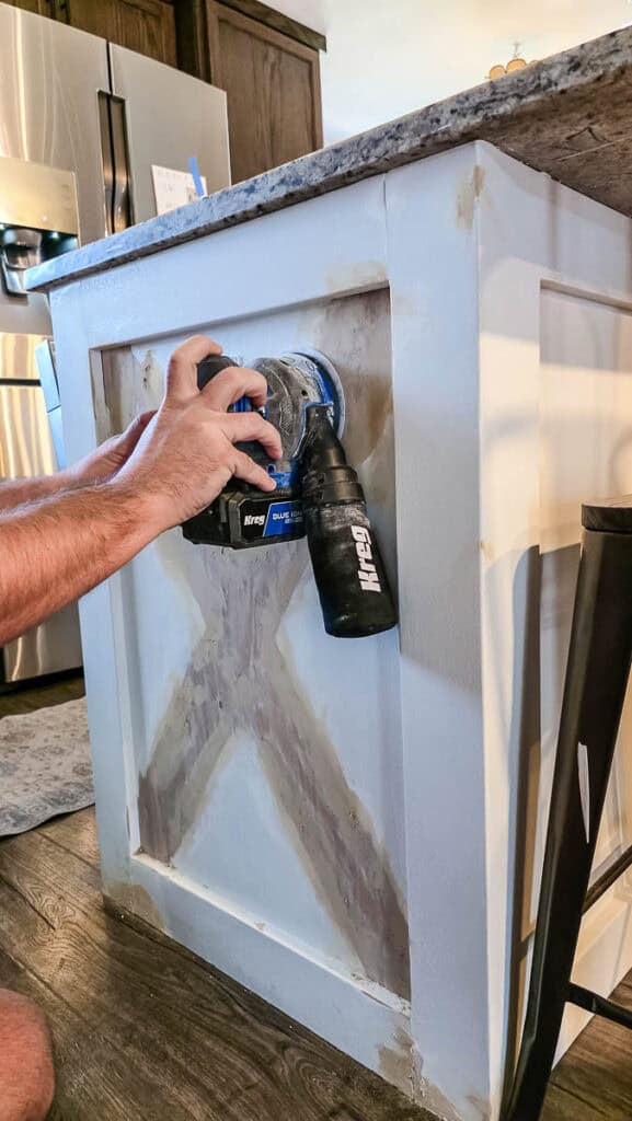 Person using an orbital sander to smooth repaired areas of a kitchen island after applying wood filler, preparing the surface for primer and paint.