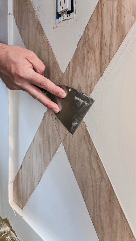 Hand using a putty knife to scrape loose paint from a kitchen island with decorative X-pattern trim, preparing the surface for sanding before painting.