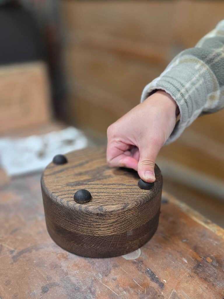 A person attaches protective rubber feet to the bottom of a stained wooden cup holder, completing the final assembly of a handmade DIY disposable cup holder for parties.