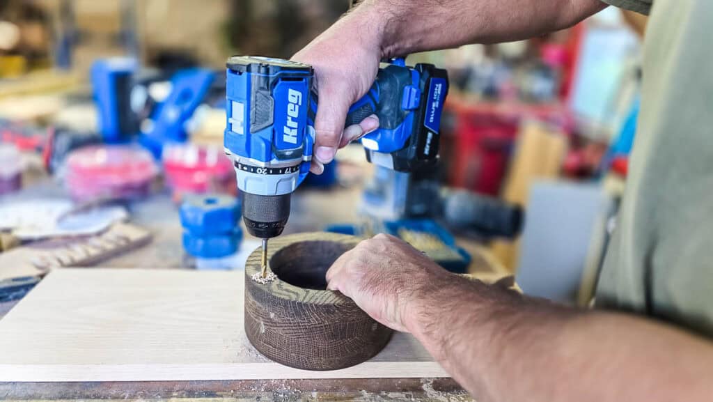 A woodworker drills a small marker hole into a wooden cup holder using a cordless drill during the final steps of building a DIY disposable cup and napkin holder for parties.
