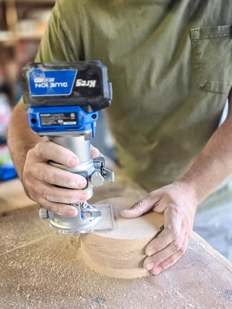 A woodworker uses a trim router to round over the outer edge of a wooden cup holder, creating smooth, finished edges for a DIY disposable cup and napkin holder.