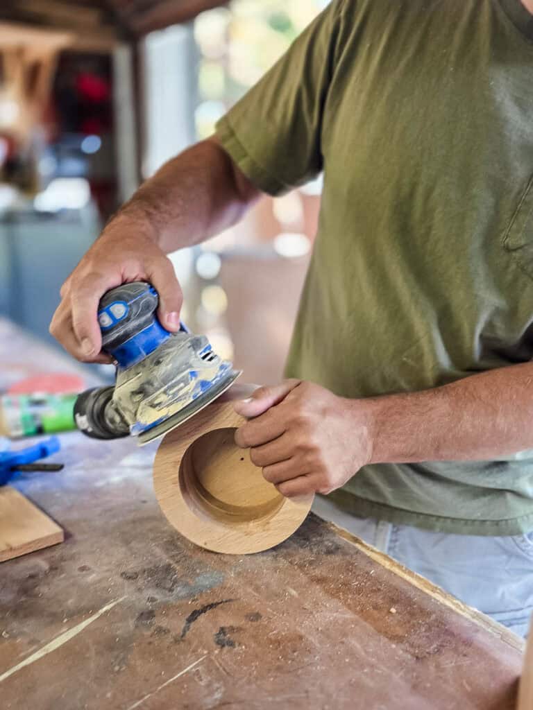 A woodworker uses an orbital sander to smooth the edges and surface of a wooden cup holder, preparing the DIY project for finishing.