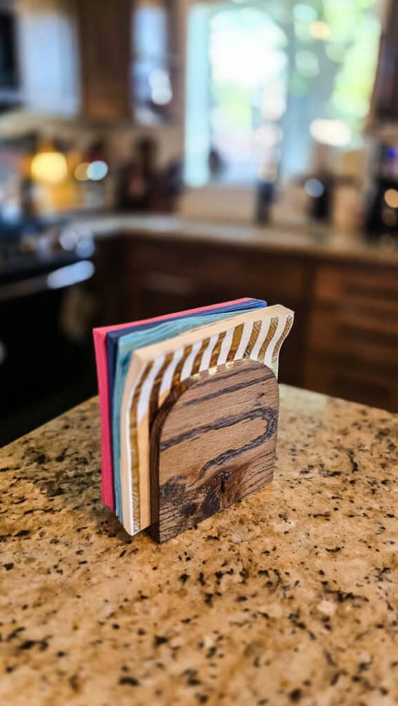 A finished handmade wooden napkin holder neatly organizes colorful disposable paper napkins on a kitchen countertop, showcasing a stylish and functional DIY party accessory.