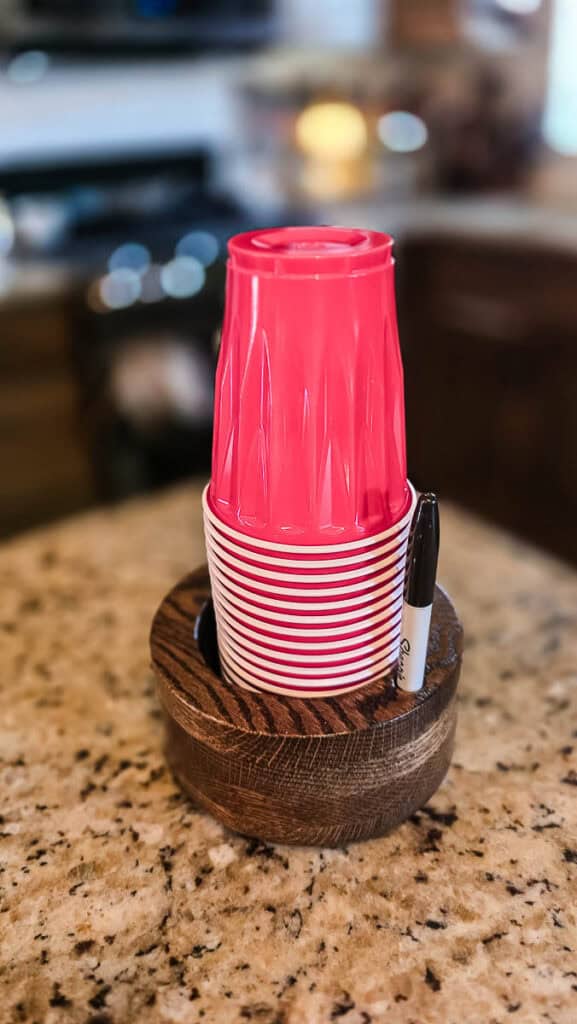 A finished DIY wooden disposable cup holder holds a stack of red plastic party cups and features a built-in marker holder for labeling drinks, displayed on a kitchen countertop.