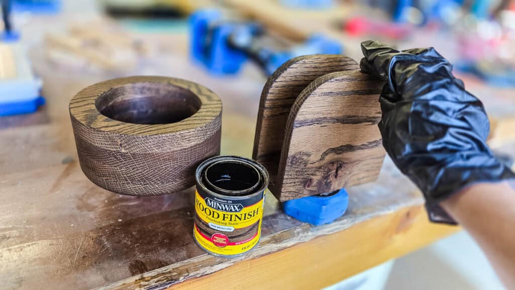 A gloved hand applies dark wood stain to a handmade wooden napkin holder beside a matching wooden cup holder, giving the DIY party organizers a smooth, rich finish.