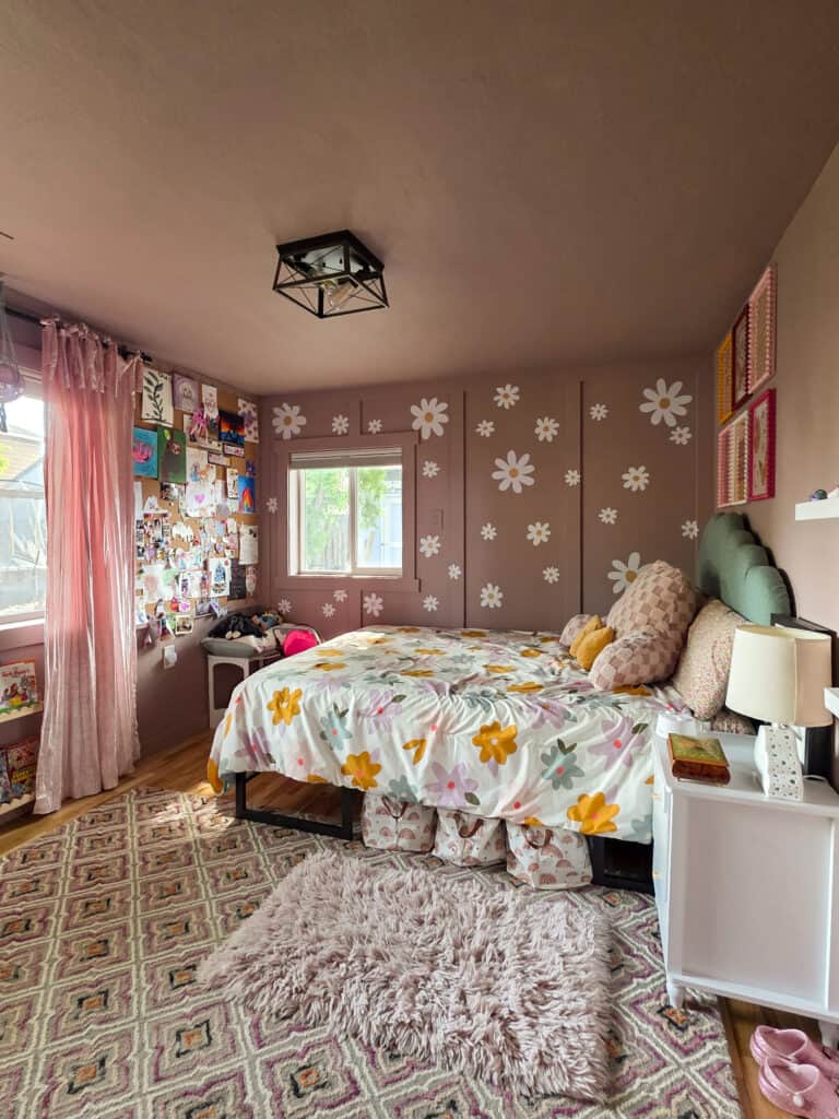 Wide view of the completed girl's pink bedroom featuring blush walls with white daisy decals, floral bedding, a scalloped green headboard, blush velvet curtains, a memory board gallery wall, patterned rugs, and cozy retro-inspired décor.