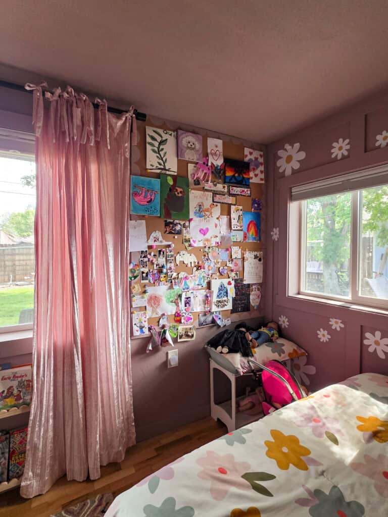 A cozy corner of a girl's pink bedroom featuring blush velvet curtains, a large inspiration board filled with children's artwork, photos, and keepsakes, plus soft daisy wall accents beside a sunny window.