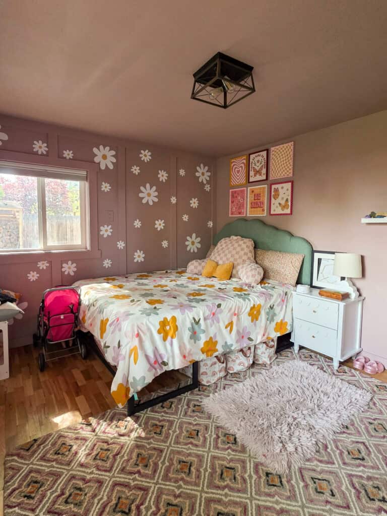 Wide view of a cozy girl's pink bedroom featuring daisy-patterned accent walls, a sage green scalloped headboard, floral bedding, colorful framed artwork, a white nightstand, and a soft shag rug.