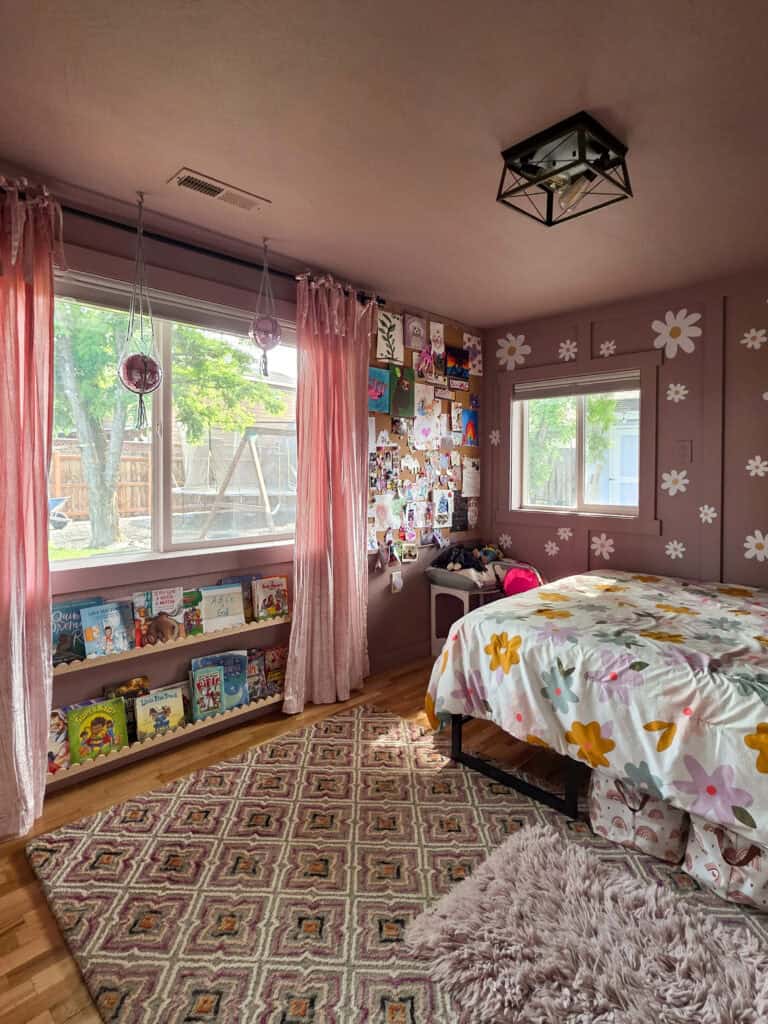 Wide view of a completed girl's pink bedroom featuring blush walls with white daisy decals, sheer pink curtains, floral bedding, a corkboard gallery wall, built-in book ledges, hanging planters, and a cozy retro-inspired reading corner.