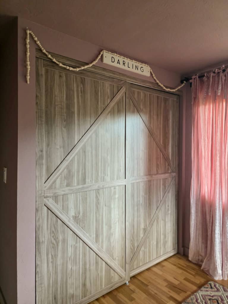 Light wood sliding barn-style closet doors in a girl's pink bedroom, decorated with a "Darling" sign, a wooden bead garland, and soft blush velvet curtains for a cozy retro-inspired look.