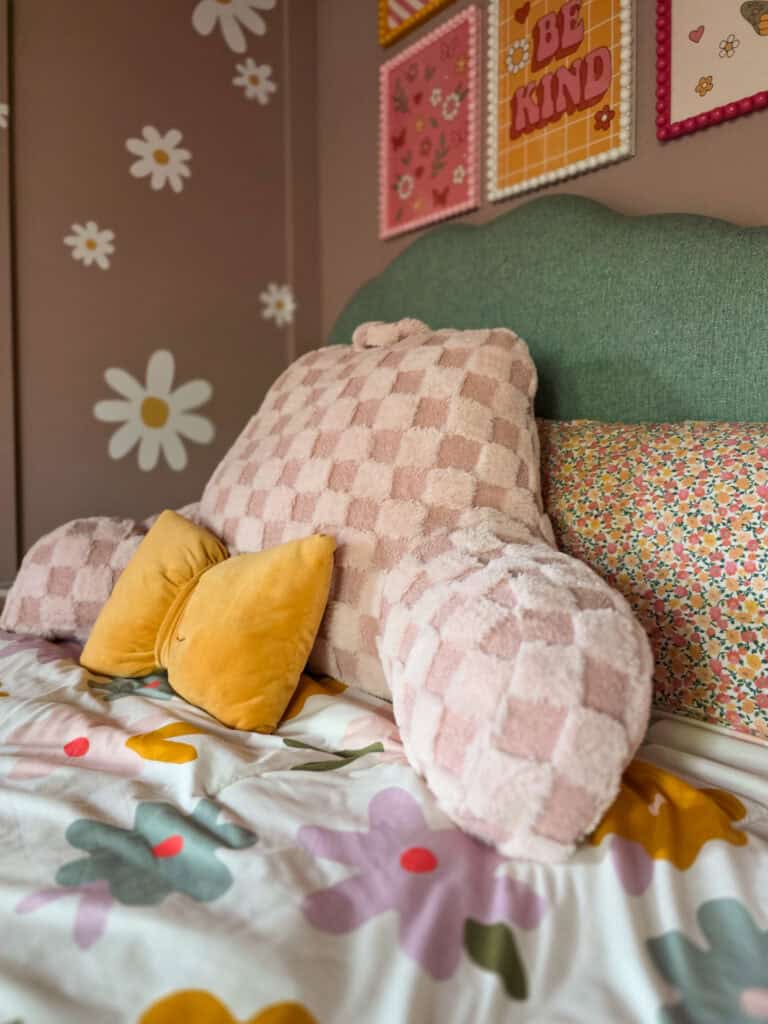 Close-up of a cozy girl's bed styled with a scalloped green upholstered headboard, blush pink checkered pillows, floral bedding, cheerful yellow accent pillows, and blush walls decorated with white daisy decals.