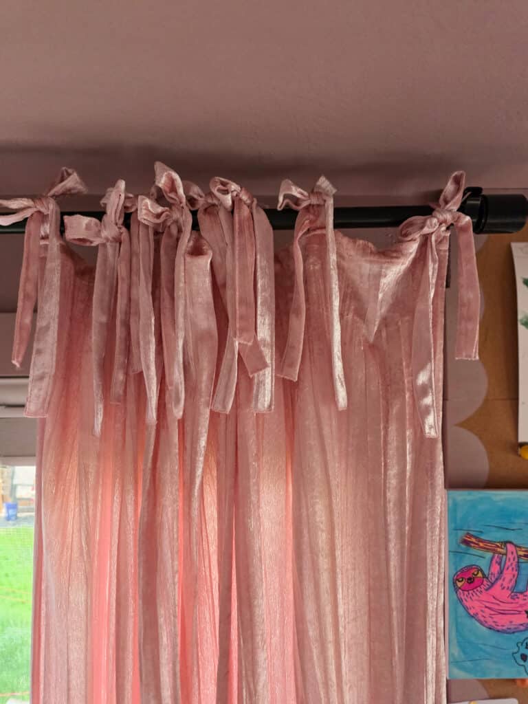 Close-up of soft blush pink velvet curtains with decorative tie-top bows hanging from a black curtain rod, adding a whimsical and cozy touch to a girl's retro-inspired bedroom.